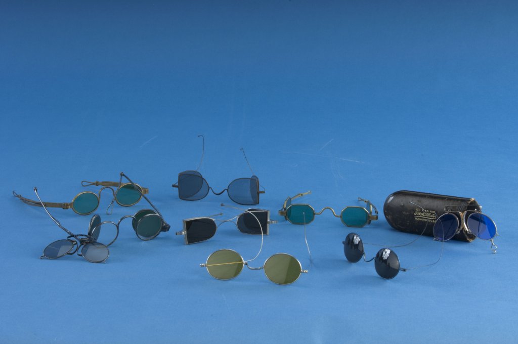 Nine spectacles variously tinted blue, green, and yellow, and a dark leather eyeglass case. 